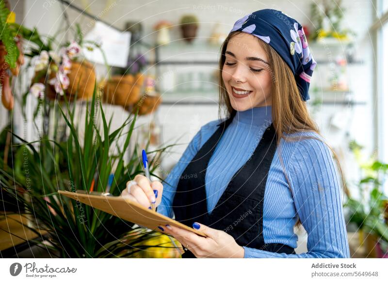 A young happy woman with a headscarf is inspecting a fern in a plant shop while holding a clipboard florist inventory check apron turtleneck indoors botanical