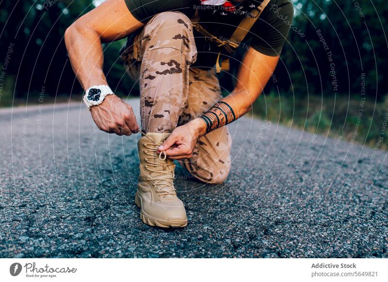 Crop anonymous Mature male commando tying shoelace while kneeling on road soldier pant blur military camouflage faceless wristwatch blurred background