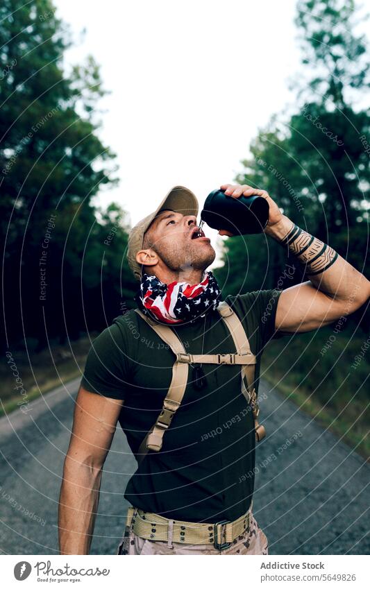 Mature army soldier drinking water while standing on road against sky man commando bottle quenching thirsty marine green cap blurred background military ranger