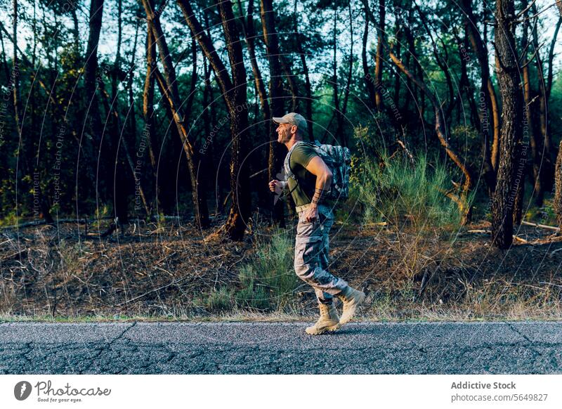 Full body of Smiling soldier training while jogging by plants and trees man commando marine running backpack road fitness uniform roadside camouflage side view