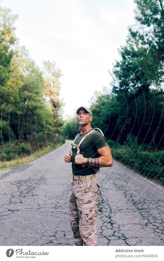 Army man in uniform with backpack and cap standing on road against trees in forest soldier confident camouflage pant green looking away army military warrior