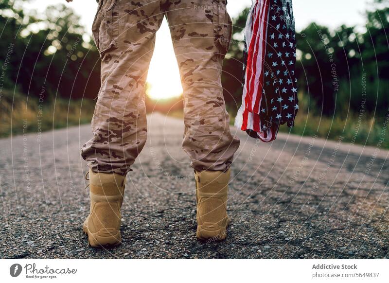 Crop legs of anonymous warrior with flag standing on road during sunset man commando american camouflage boot uniform frontline faceless blurred background