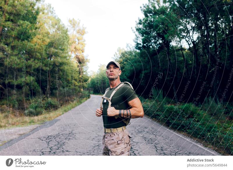Army man in uniform with backpack and cap standing on road against trees in forest soldier confident camouflage pant green looking away army military warrior
