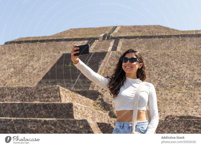 Positive stylish traveler taking selfie during trip to tourist site in Mexico woman sightseeing mexico sunglasses pyramid smartphone casual vacation building