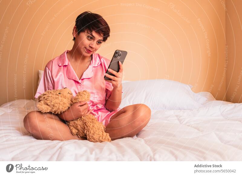 Smiling young female with soft teddy bear browsing smartphone in bedroom woman using smile at home social media happy internet nightwear cellphone positive