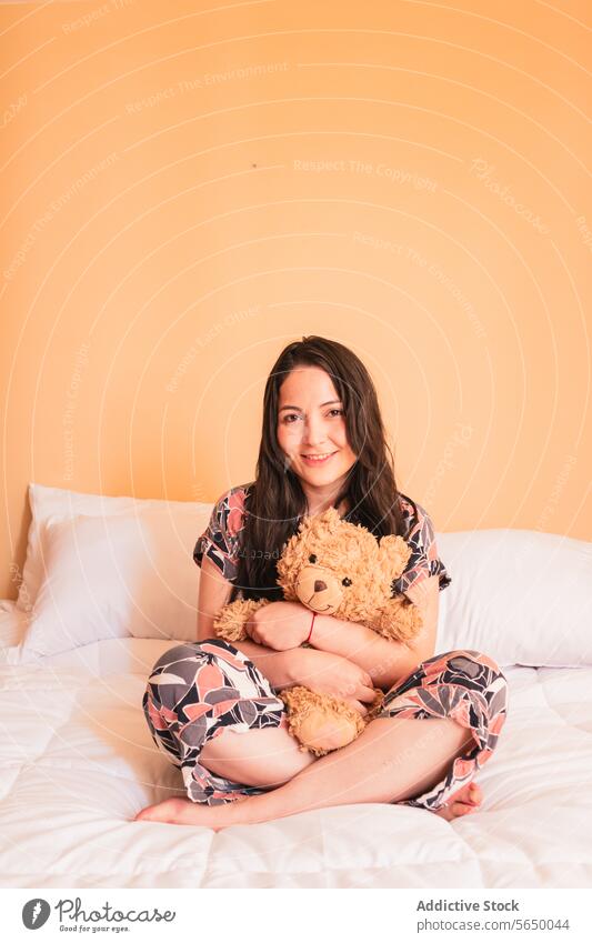 Young woman sitting on bed with teddy bear toy hug smile plush cozy bedroom female young comfort happy pajama sleepwear relax lady peaceful legs crossed calm