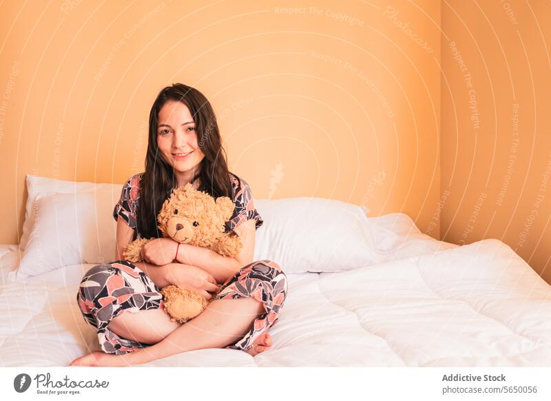 Young woman sitting on bed with teddy bear toy hug smile plush cozy bedroom female young comfort happy pajama sleepwear relax lady peaceful legs crossed calm