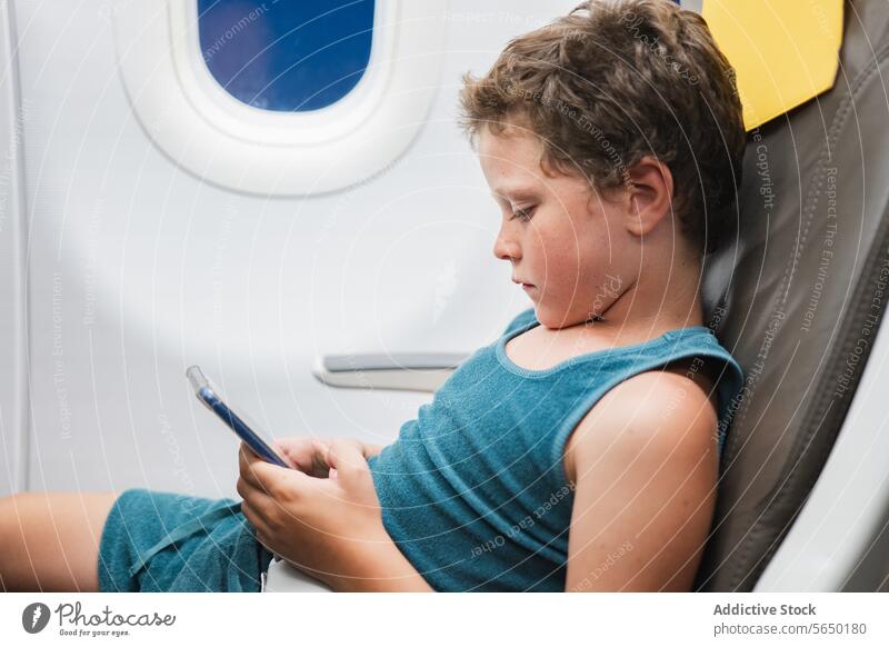 Young Boy Engrossed in Tablet on Plane boy child airplane seat tablet cellphone screen technology travel flight window young passenger digital device
