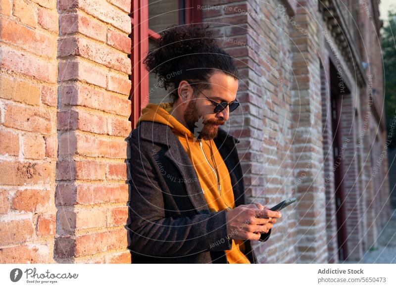 Stylish man with smartphone in urban setting street sunset style fashion city young adult male curly hair topknot texting casual wear sunglasses facial hair
