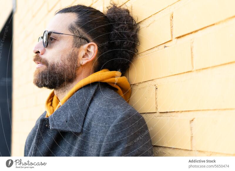 Modern man with sunglasses leaning against a yellow brick wall style modern gray coat scarf thoughtful day sunny fashion hipster urban outdoor casual hairstyle