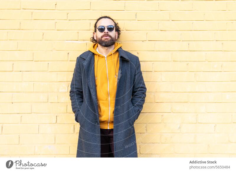 Casual man leaning against a yellow brick wall sunglasses hoodie urban street style confidence standing casual male fashion relaxed pose outdoor texture