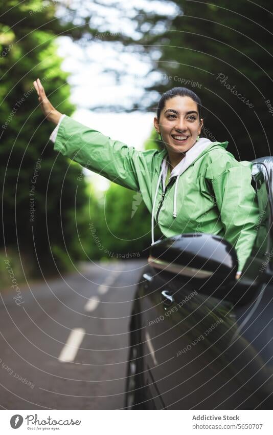 Happy young woman with arms outstretched looking away and leaning out of car window while enjoying road trip in forest carefree happy journey smile raincoat
