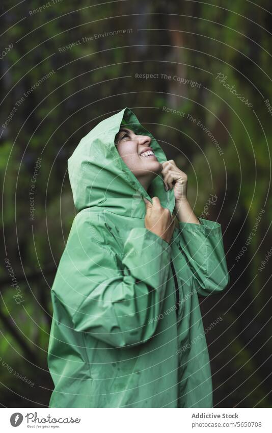 Smiling young woman wearing green raincoat with hoodie looking up while hiking by cliff covered in moss during vacation smiling trail forest smile covering