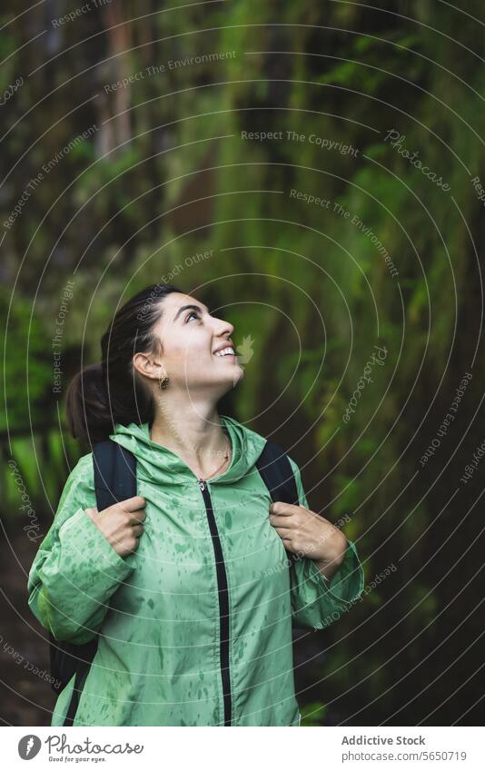 Smiling young woman wearing green raincoat looking up while hiking by cliff covered in moss during vacation smiling trail forest smile covering walking