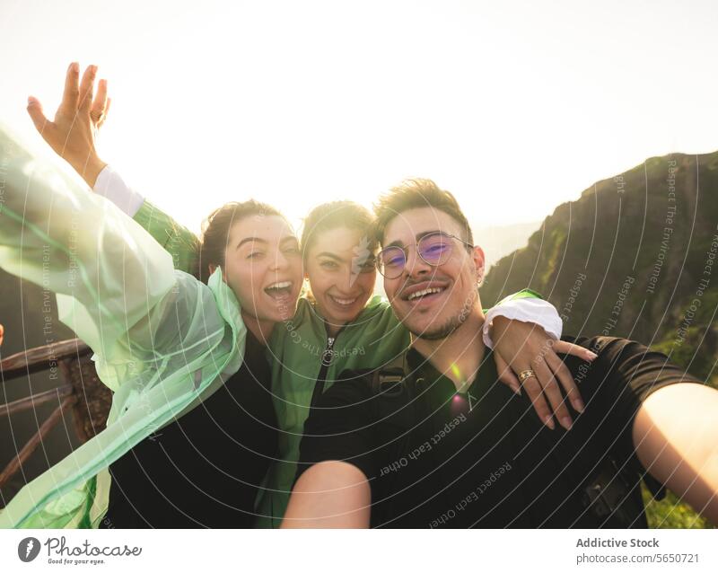 Portrait of cheerful Caucasian active young friends taking selfie looking at camera on mountain peak during sunrise at weekend hiking women man portrait backlit