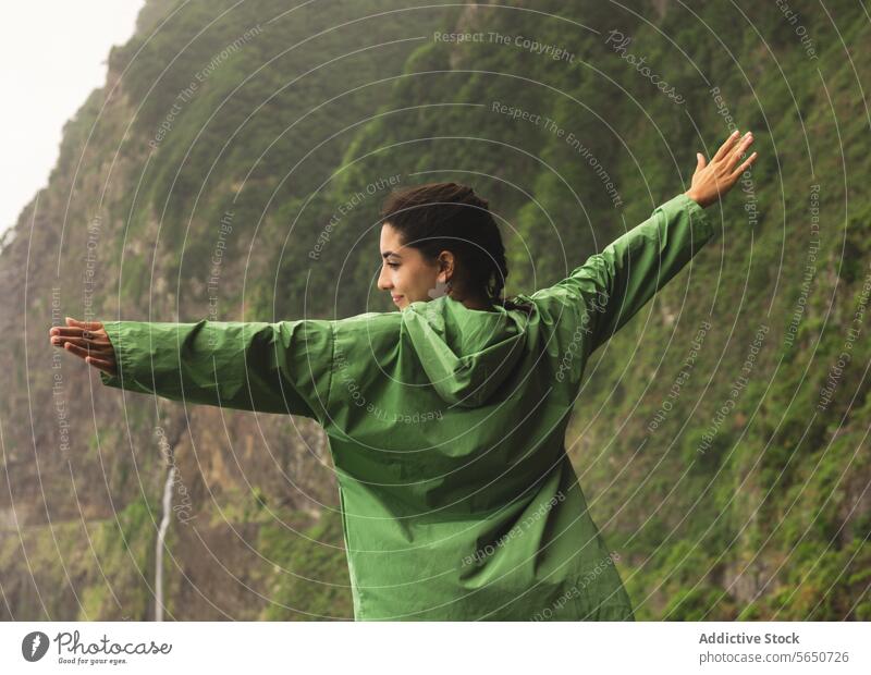 Back view of carefree young woman in green raincoat standing with arms outstretched on concrete barrier against mountains back view hiker seaside enjoy hiking