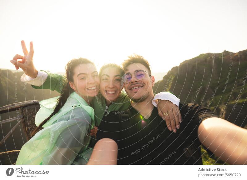 Portrait of cheerful Caucasian active young friends taking selfie looking at camera on mountain peak during sunrise at weekend hiking women man portrait backlit