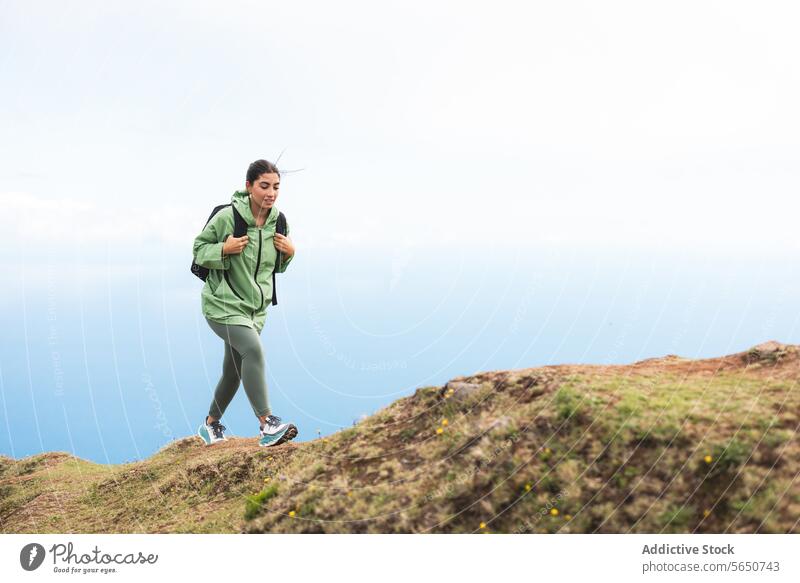 Full body side view of young female hiker in green raincoat with backpack moving up on mountain peak against sky during weekend holiday woman walking top