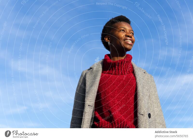 From below smiling African American female in red turtleneck sweater and gray coat walking with hands in pockets against blue sky woman stroll weekend free time