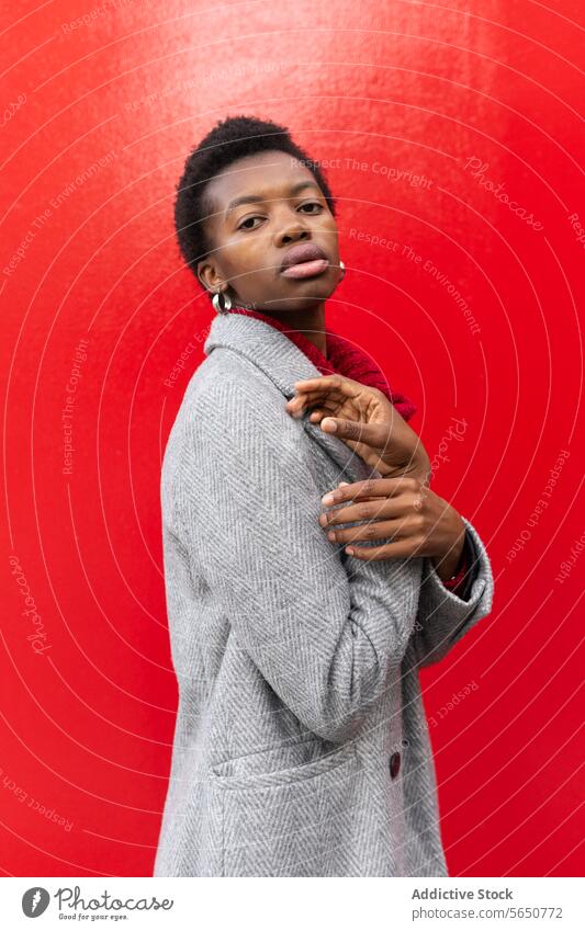 Side view of emotionless African American woman with short hair in warm jacket and earrings looking at camera standing on red background style individuality