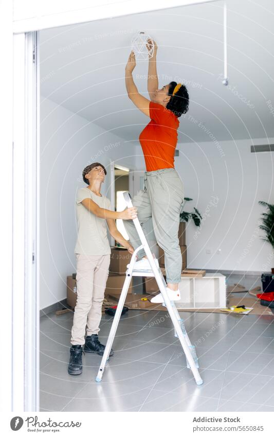 Mom on ladder fixing lamp and son holding ladder and looking up mom relocation hang woman mother help plant relocate together apartment casual new improve home