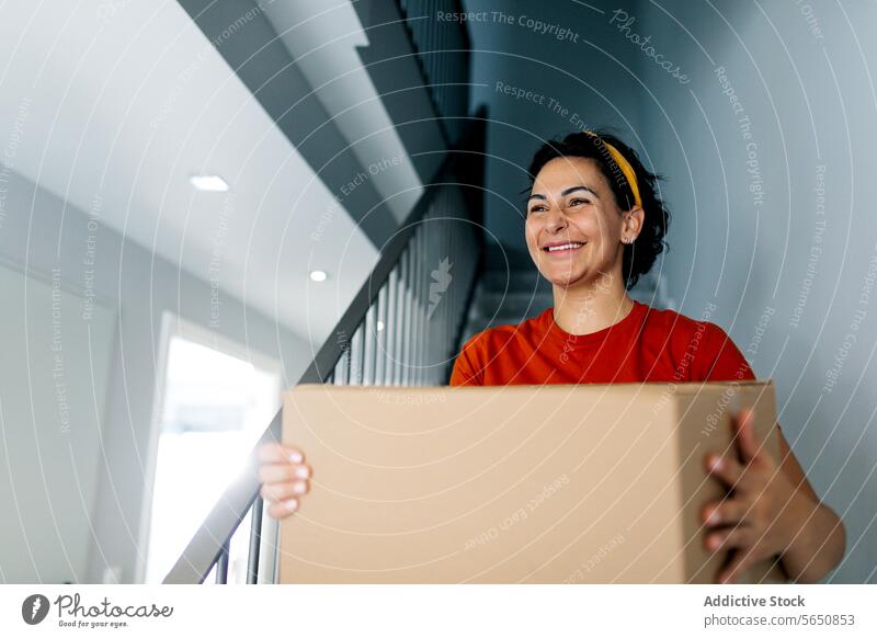 Smiling woman with box on stairs in new apartment cardboard carry relocate move in home staircase cheerful belonging stairway casual smile female estate