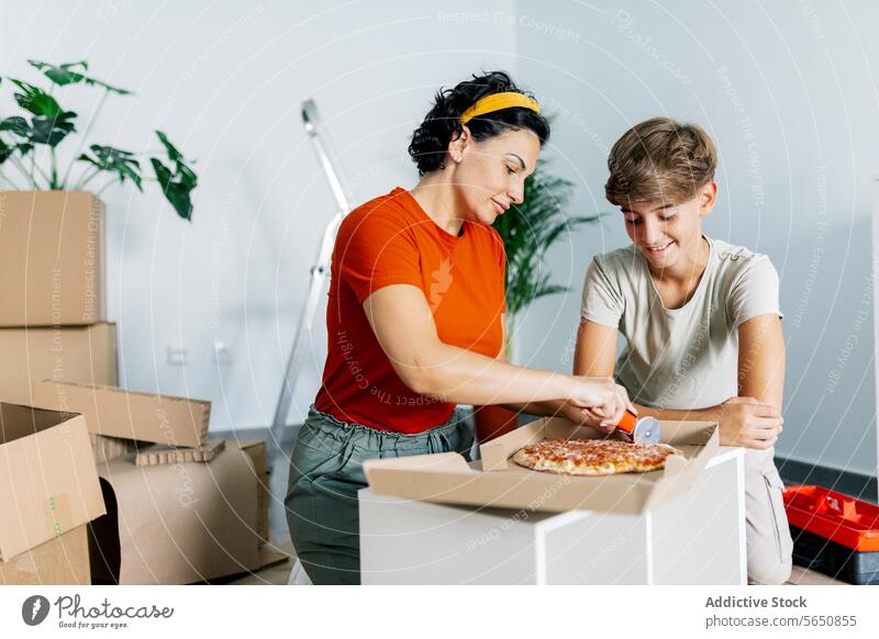 Happy mother slicing pizza with cutter and son watching with delight appetizing unpack belonging move in new flat apartment home kitchen together relocate smile