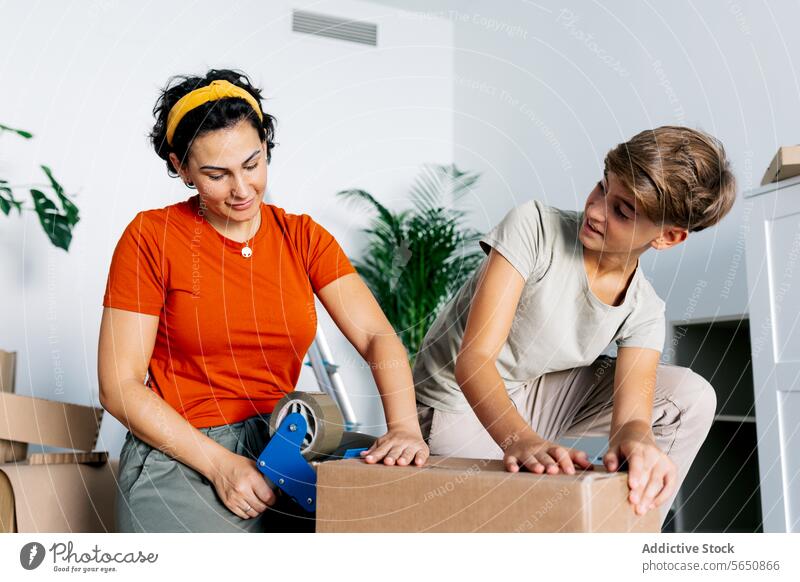 Focused mother in red top packaging box with tape dispenser with son help apartment new package teenage move relocate home boy together indoors casual house