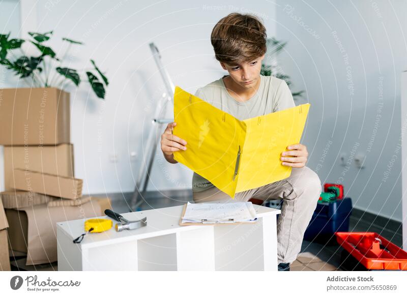 Focused boy kneeling and reading notes on yellow clipboard instruction assemble furniture check relocate teenage focus examine home serious casual concentrate