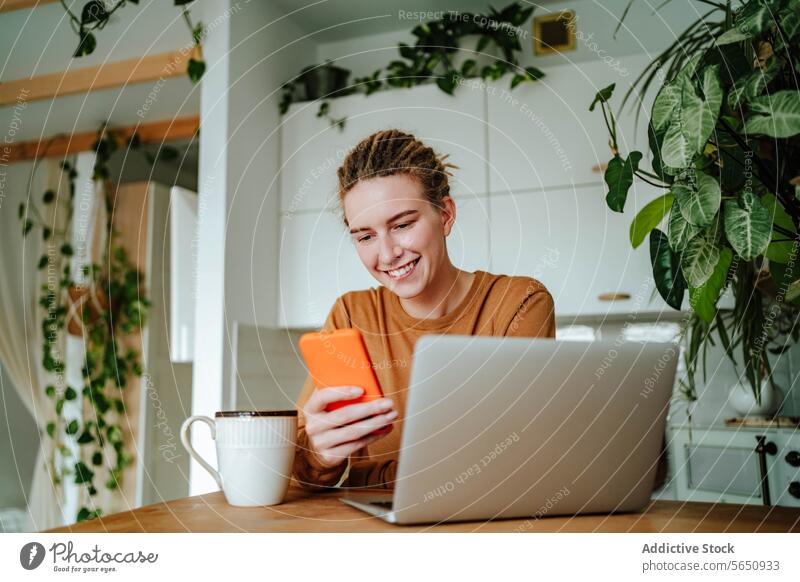 Smiling young woman using smartphone at kitchen table with laptop in daylight happy smile text message online at home female sit dreadlocks browsing surfing