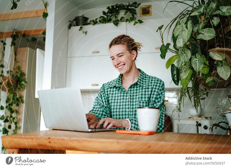 Happy young woman working on laptop at table in decorated kitchen at home freelance remote startup online female sit checkered shirt gadget device smile happy