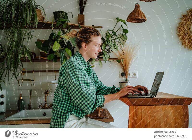 Happy young woman working on laptop at table in decorated kitchen at home freelance remote startup online female sit checkered shirt gadget device smile happy