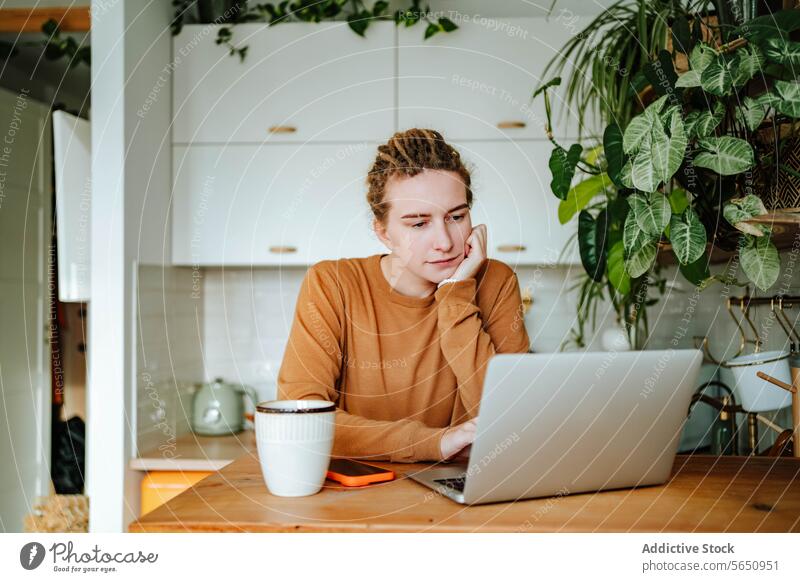 Young woman working on laptop at kitchen table with smartphone coffee mug in daylight thoughtful lean on hand cabinet recipe at home female young dreadlocks