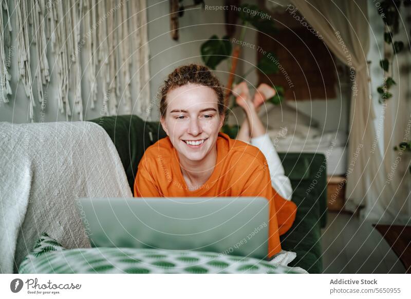 Smiling woman working on laptop while lying on sofa at home using online remote freelance smile browsing internet happy self isolation couch connection comfort