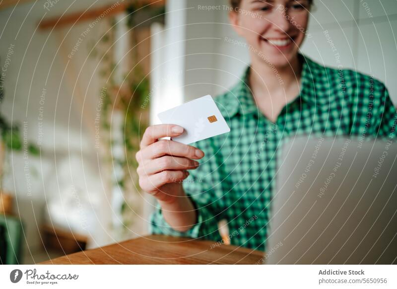 Blurred happy young woman sitting with credit card near laptop at table in daylight purchase online smile shop glad at home female positive device gadget