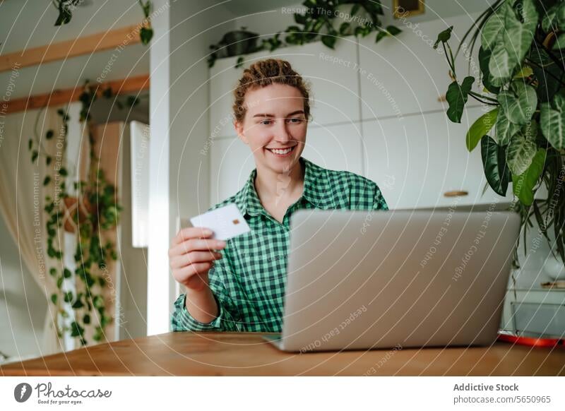 young woman sitting with credit card near laptop at table in daylight purchase online smile shop happy glad at home female positive device gadget shopper