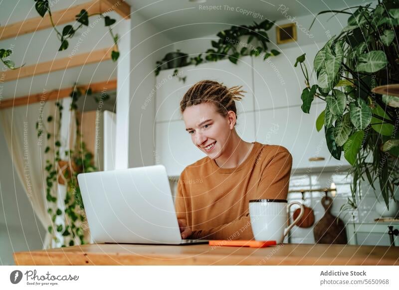 Happy Young woman working on laptop at kitchen table with smartphone and coffee mug in daylight cabinet at home female young dreadlocks gadget device mobile