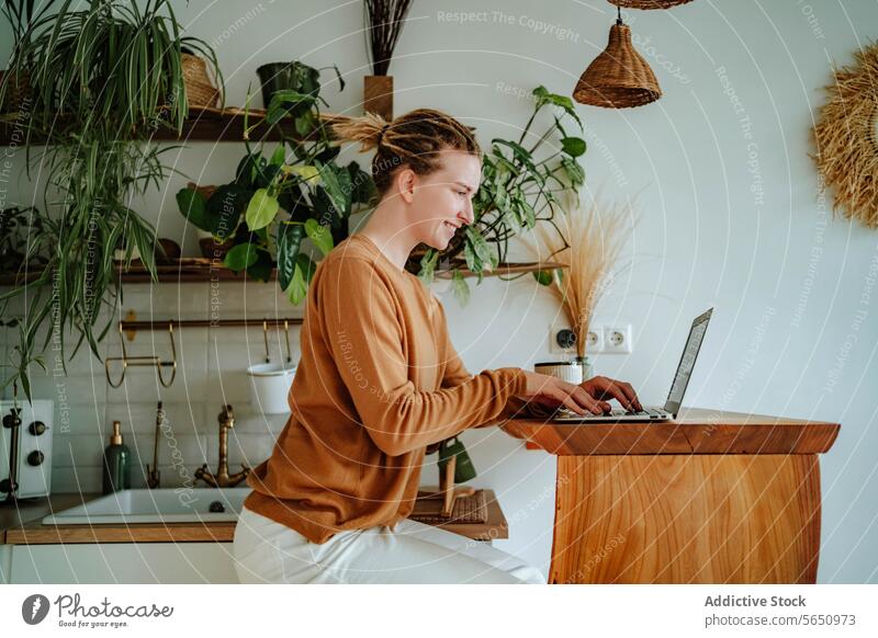Happy Young woman working on laptop at kitchen table with coffee mug in daylight cabinet at home female young dreadlocks gadget device online netbook browsing
