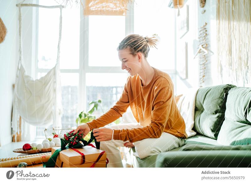 Side view if Young woman preparing present box at home while sitting on sofa gift smile surprise happy joy casual comfort wrap celebrate couch positive glad