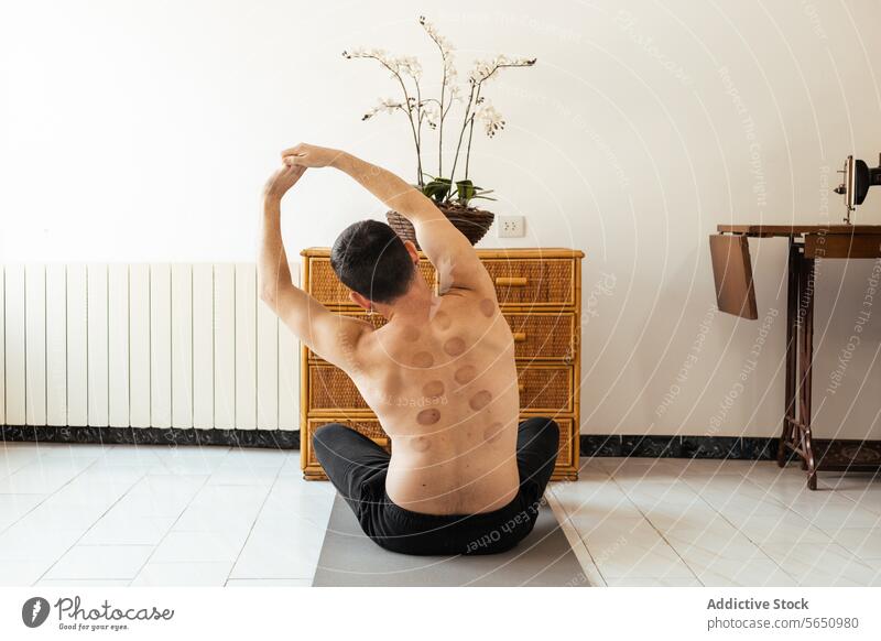 Young male sitting on floor and stretching hands up during rehabilitation training man hand up shirtless home workout mat exercise warm up practice wellness