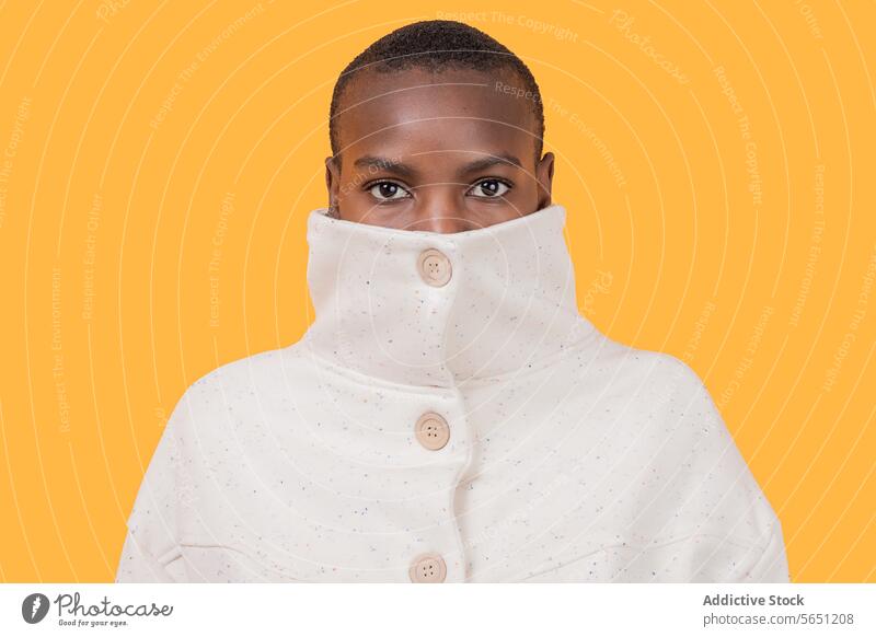 Black woman in turtleneck clothing isolated on yellow background trendy apparel covering mouth face portrait short hair staring confident style glance