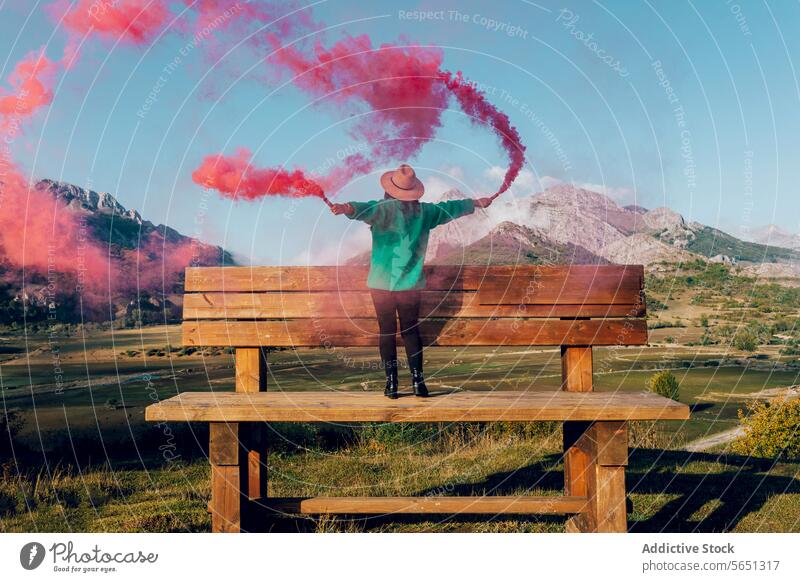 Joyful woman standing on big bench with colored smoke against mountain backdrop person smoke flare pink landscape scenic outdoor nature arms outstretched joyful