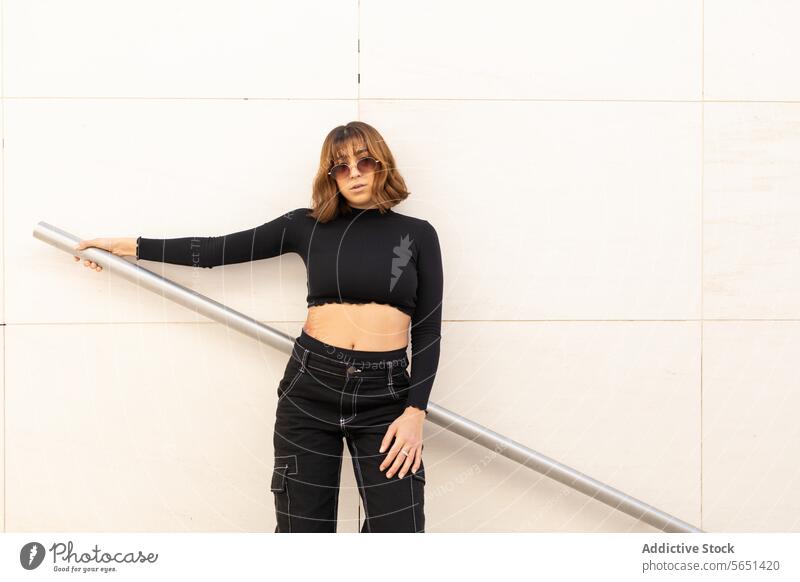 Fashionable young woman posing by a metal railing fashionable style crop top pants black silver confident neutral wall background stylish modern urban casual