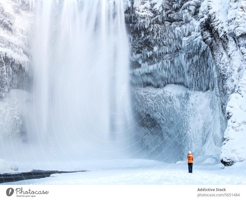 Anonymous person in an orange jacket stands in awe before the majestic icy Skógafoss waterfall in Iceland Person winter ice snow nature landscape travel cold