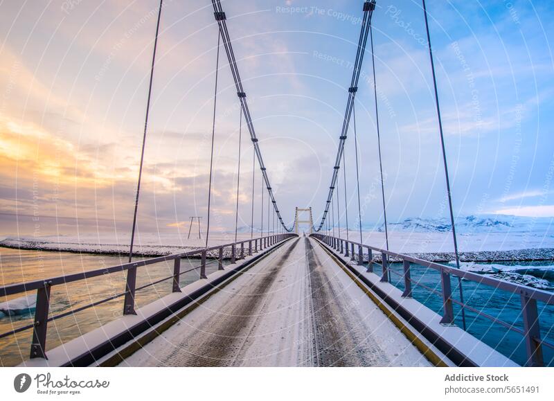 Suspension bridge with steel cables spanning a frozen river against a backdrop of snow-covered mountains at dusk suspension bridge Iceland winter landscape