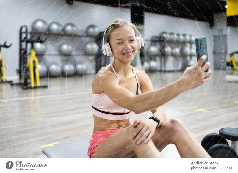 Smiling woman taking selfie at gym during workout smiling sportswear headphones smartphone fitness exercise happiness technology lifestyle health wellness