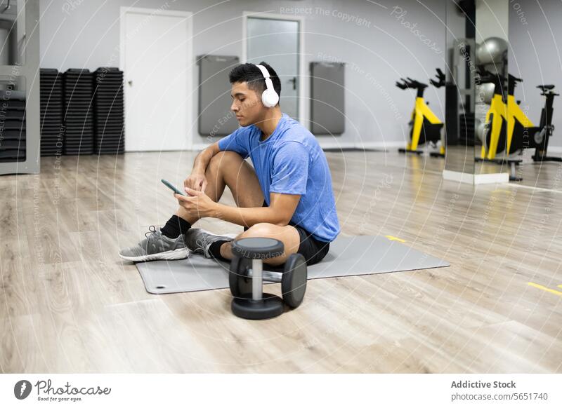 Young man using smartphone during gym break exercise equipment headphones sitting mat focused young adult fitness technology wireless communication leisure