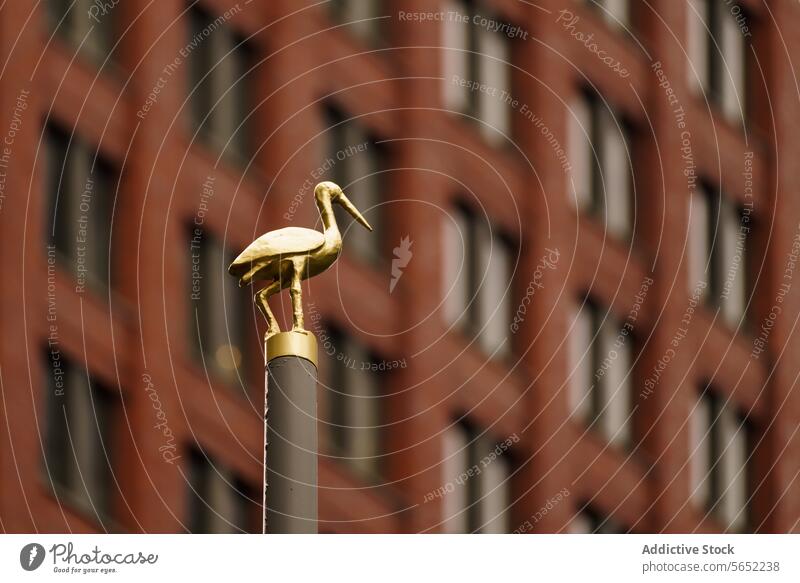 Golden stork sculpture atop a post with a blurred background of brick buildings, symbolizing The Hague golden city art installation public statue ornamental