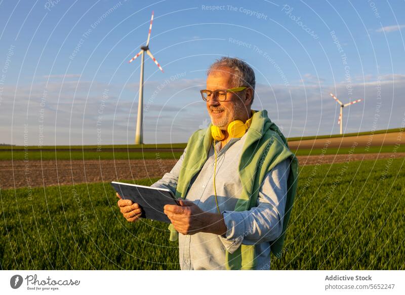 Happy senior man with headphones using digital tablet at wind farm Man Headphones Digital Tablet Windmill Farm Blue Sky Senior Using Agriculture Landscape