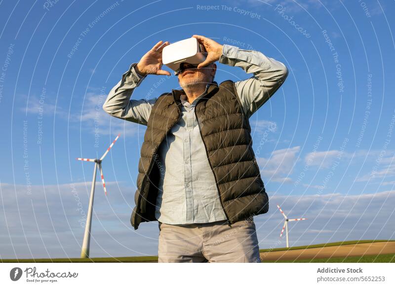 Elderly man wearing VR glasses against wind turbine and blue sky Man Glasses VR headset Windmill Gadget Senior Innovation Futuristic Stand Renewable Energy